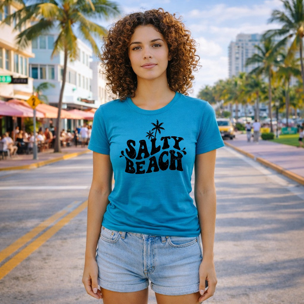 Woman wearing a blue t-shirt with 'Salty Beach' text on a street with palm trees and buildings.