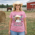 Woman wearing a pink t-shirt with 'Cow Girl Baddie' design in a rural setting with a horse and barn.